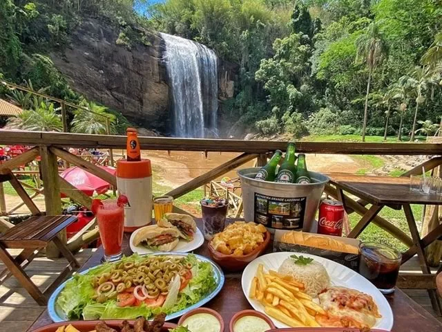Mesa repleta de petiscos e bebidas com a Cachoeira Grande ao fundo